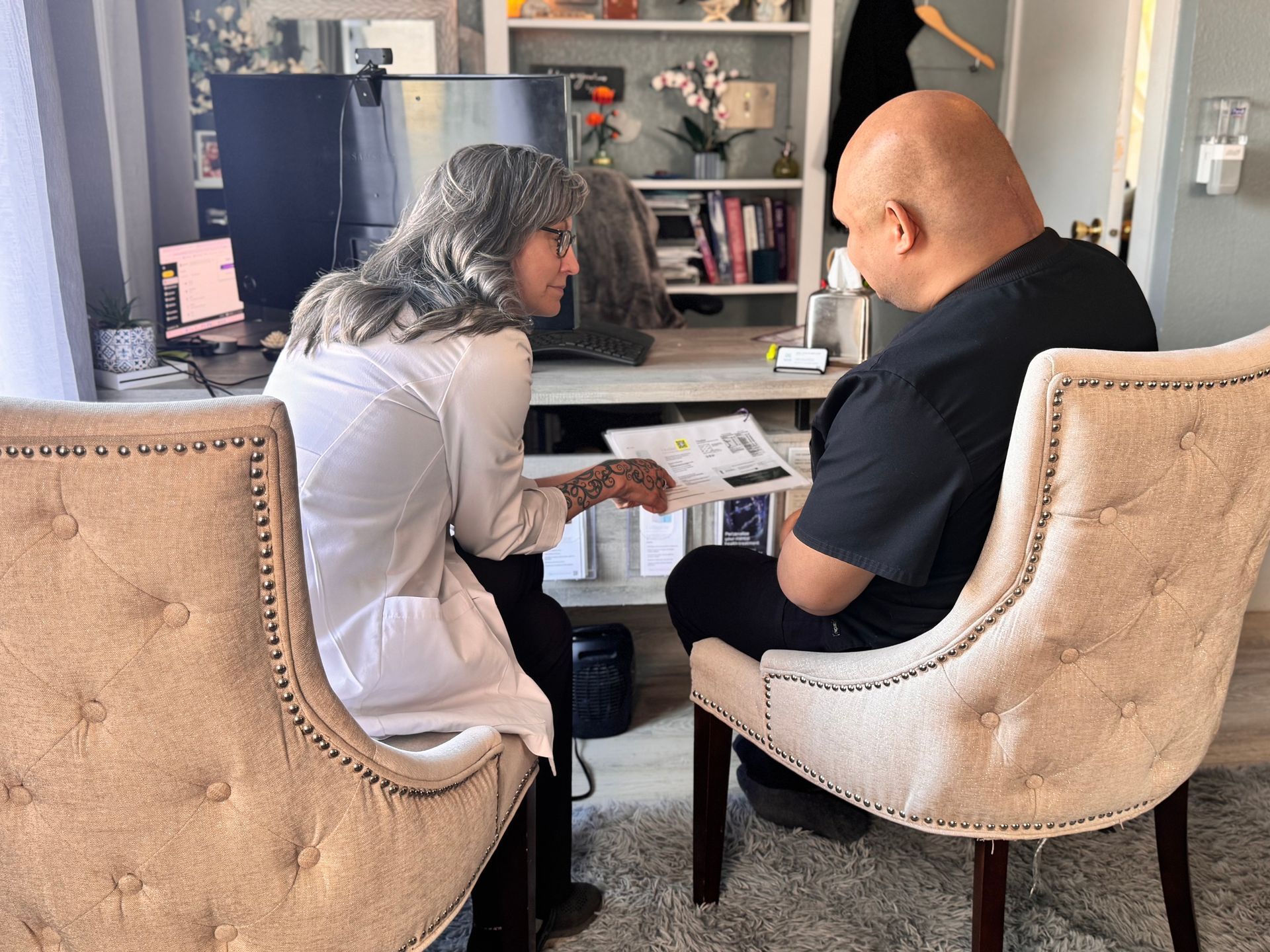 Two people sit in chairs facing each other, reviewing a document together in a home office setting.