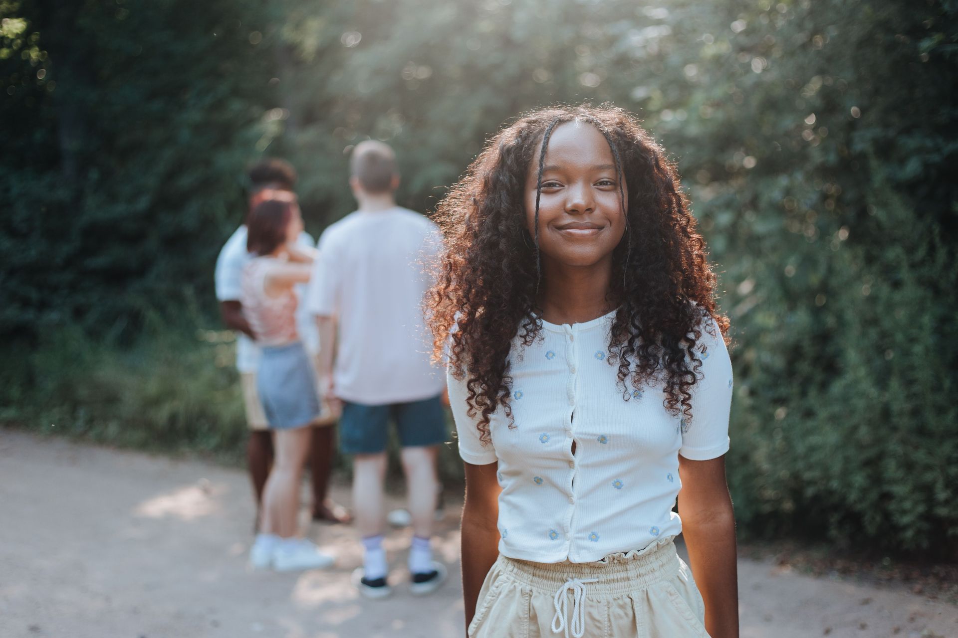 Young woman smiling on a wooded path, with two people blurred in the background.