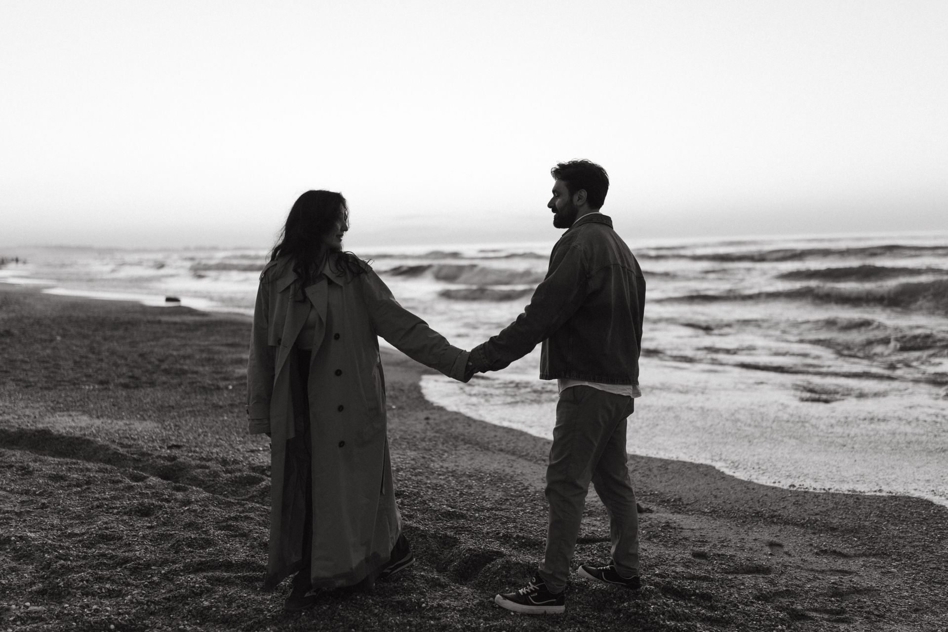 Two people holding hands on a rocky beach by the ocean, facing each other under a cloudy sky