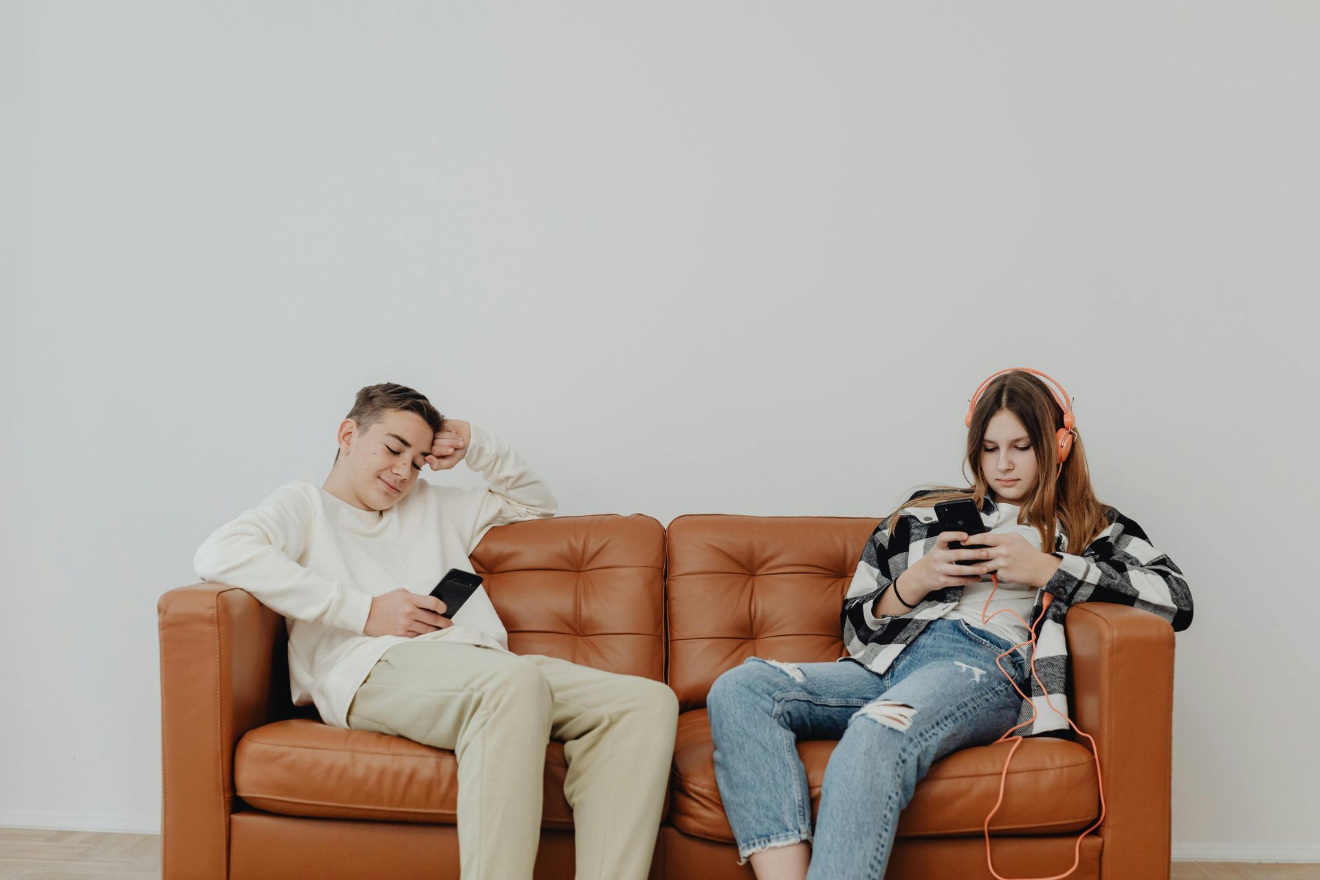 Two people on a brown couch using phones. One leans back, the other wears headphones. Plain white wall behind.