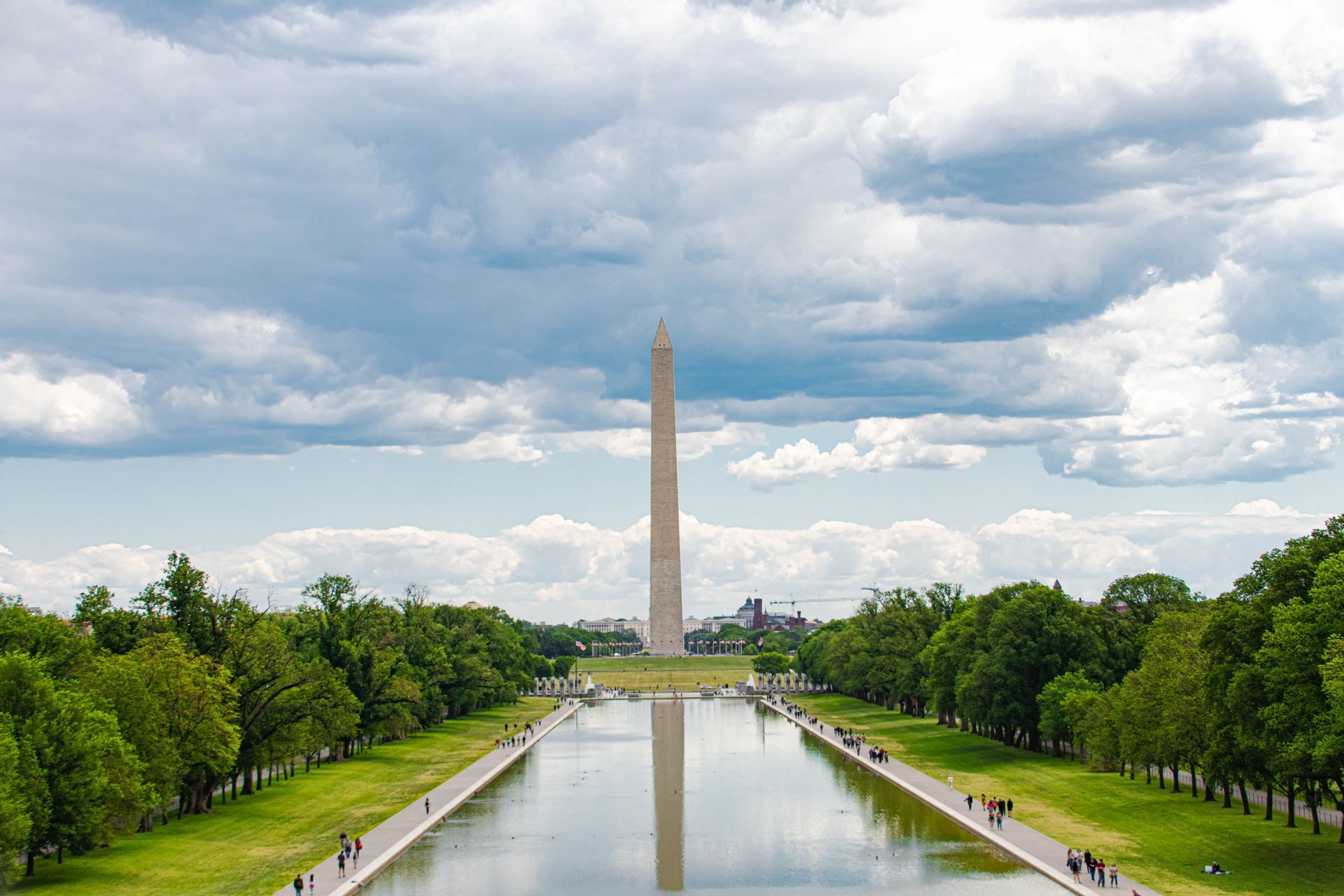 Washington Monument centered beyond Reflecting Pool, framed by green trees under cloudy sky