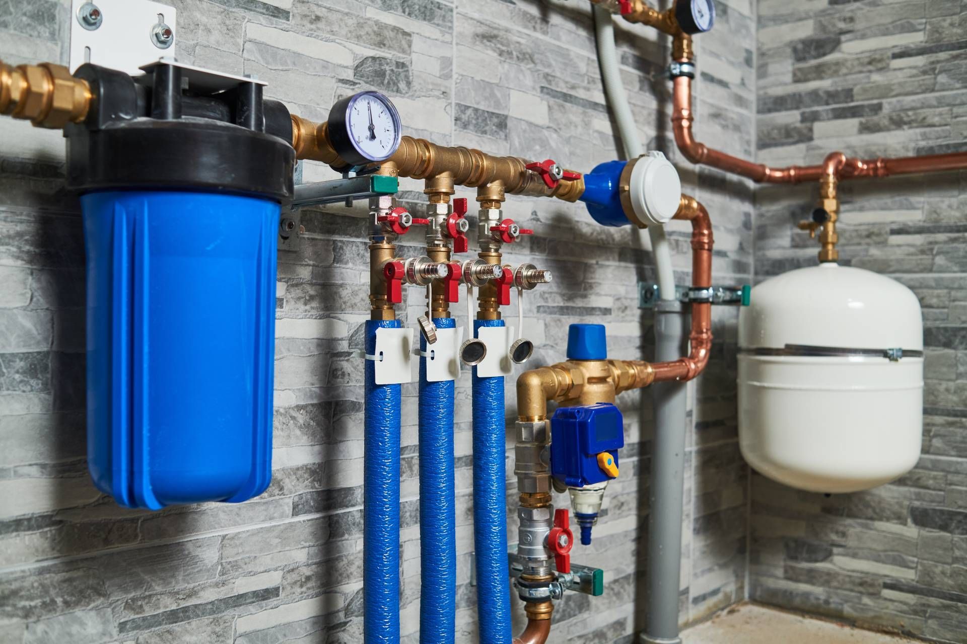Plumbing manifold with blue pipes, gauges, valves, and white expansion tank on a basement wall