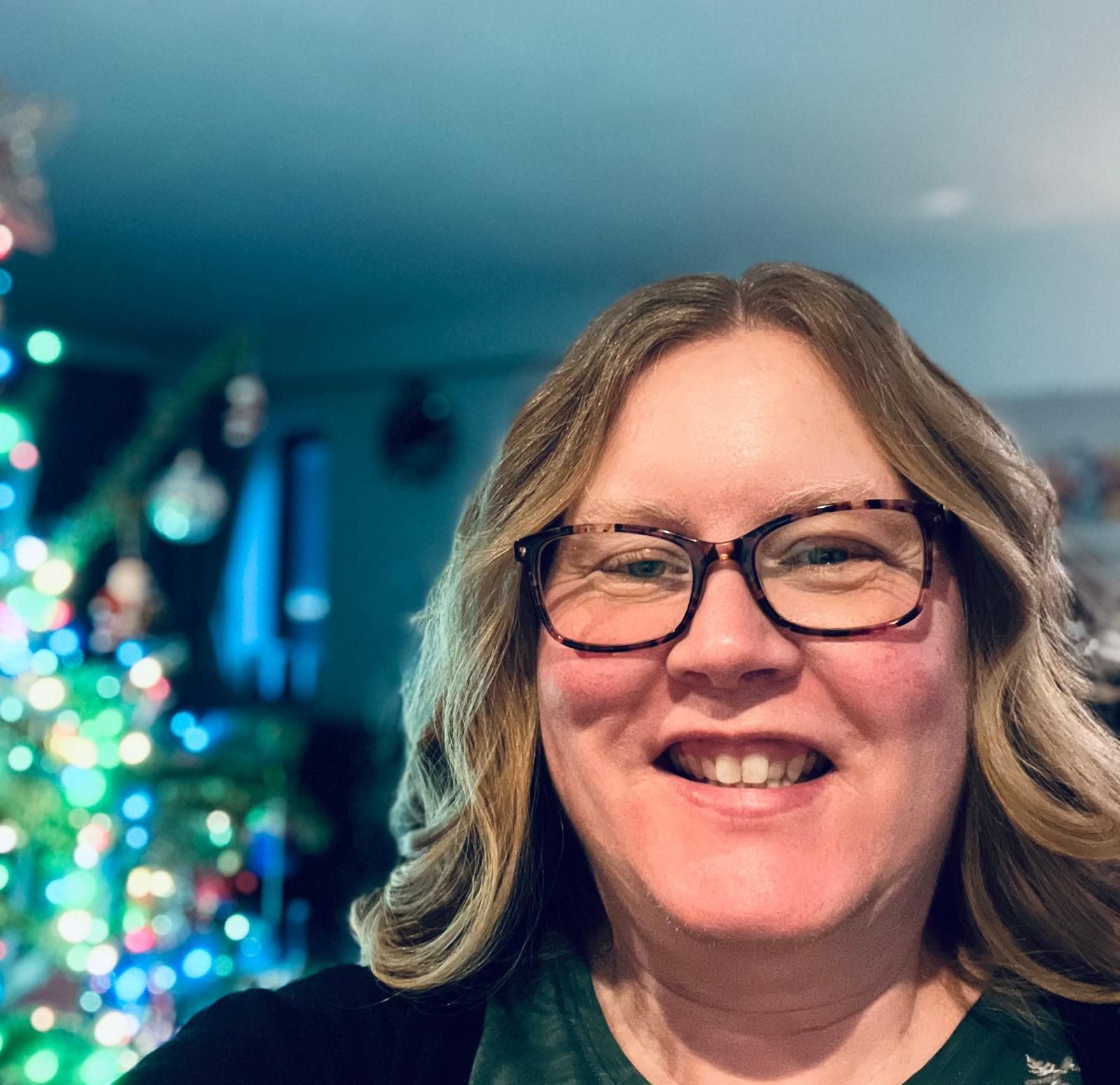 Woman with glasses smiles in front of a decorated Christmas tree with blue and green lights.