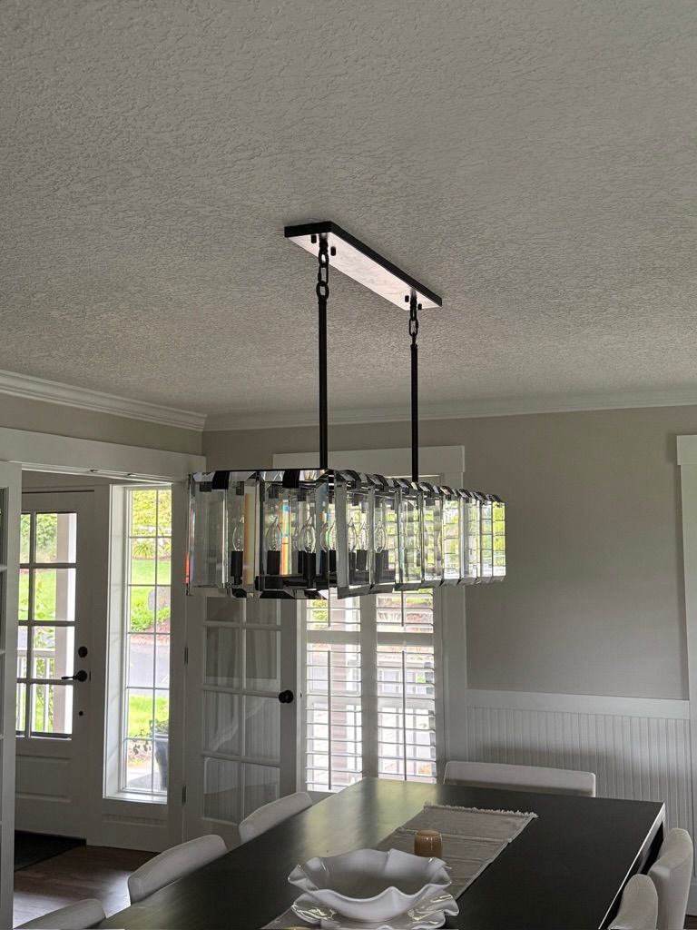 Modern dining room with a rectangular chandelier over a dark table, windows, and white chairs.