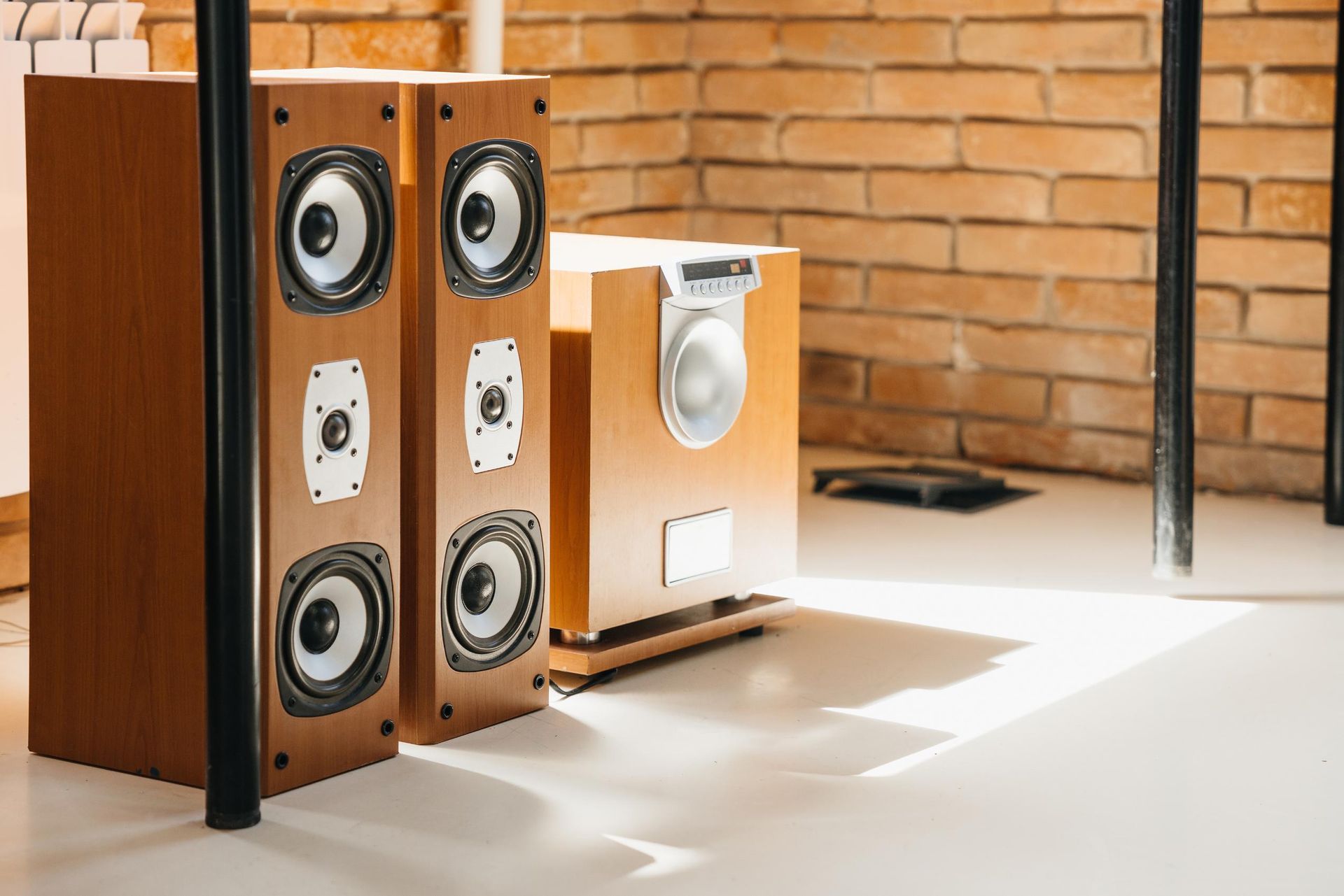 Two tall wooden speakers and a smaller speaker on a white floor next to a brick wall.