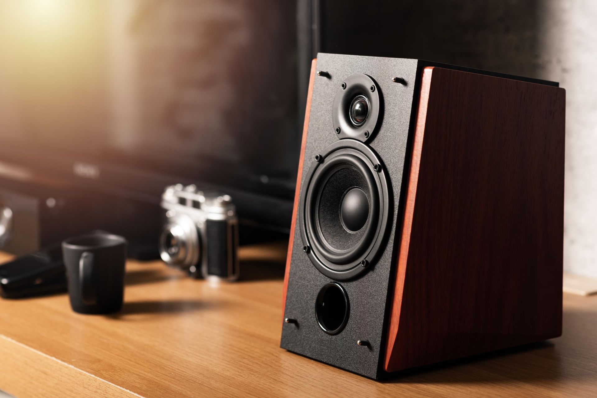 Wooden speaker on a table in front of a TV, with a black mug, camera, and remote.