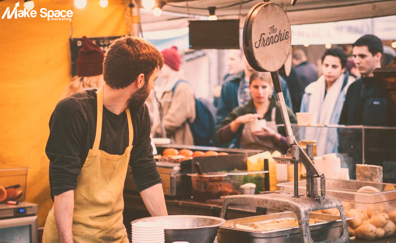 A man in an apron is standing in front of a food stand.