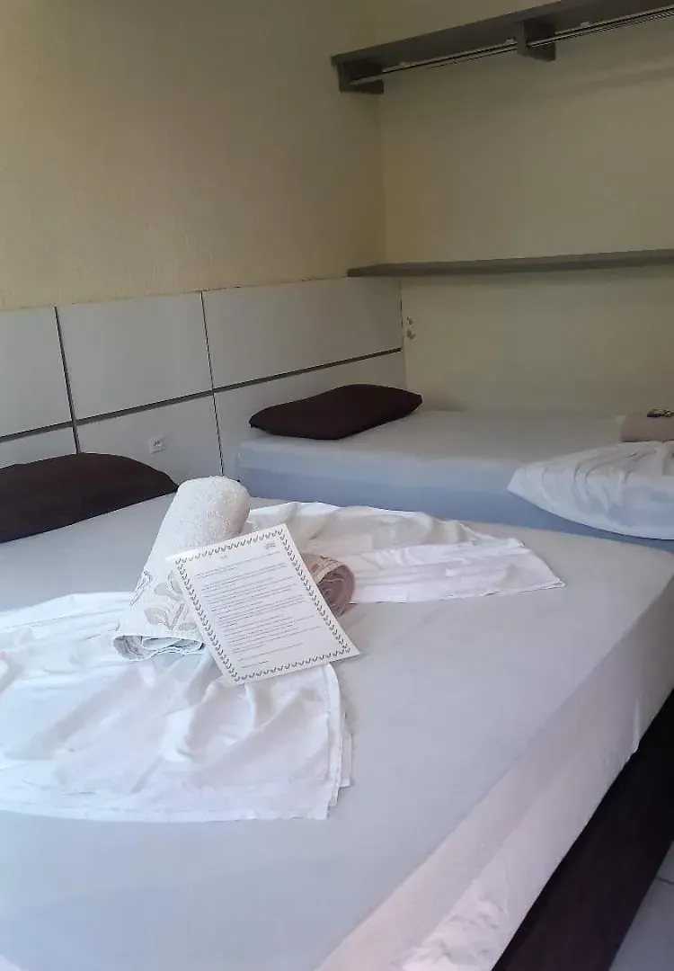 Cama de hospital com lençóis brancos, toalhas dobradas e um travesseiro marrom encostada em uma parede de azulejos.