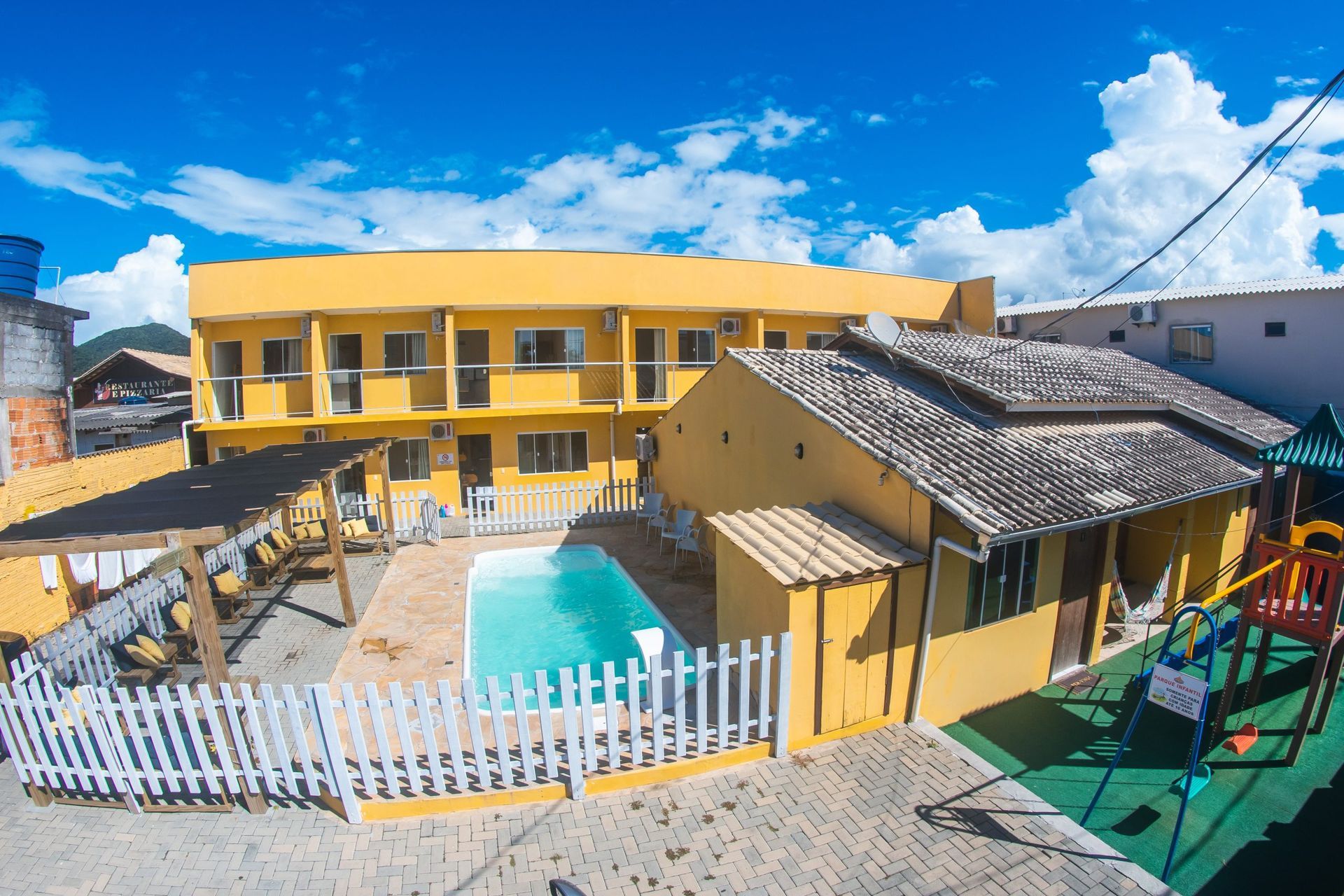 Albergue amarelo de dois andares com uma pequena piscina, cerca branca e parquinho infantil sob um céu azul brilhante.