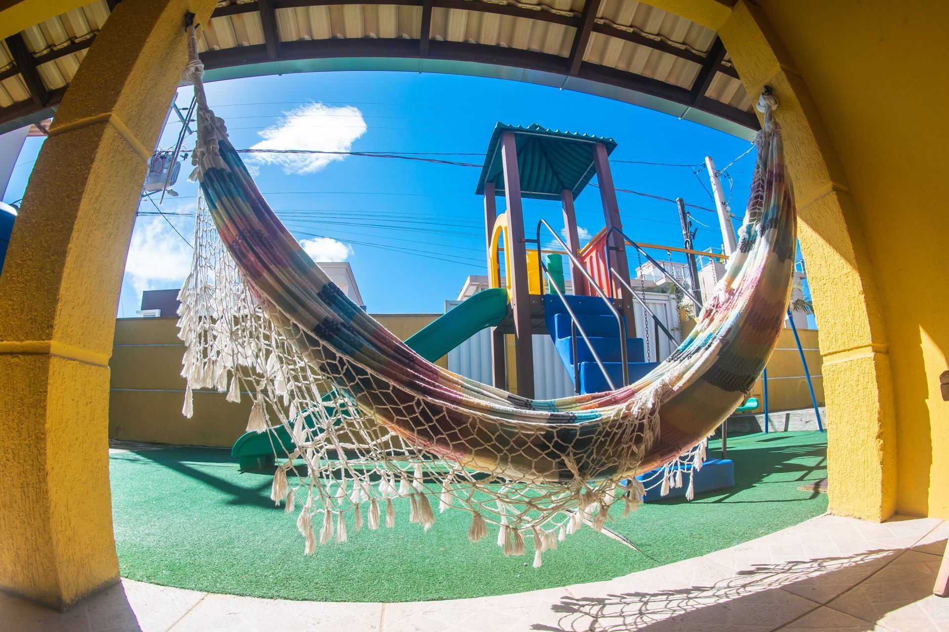 Rede suspensa sob um pátio sombreado, com um parque infantil colorido e céu azul ao fundo.