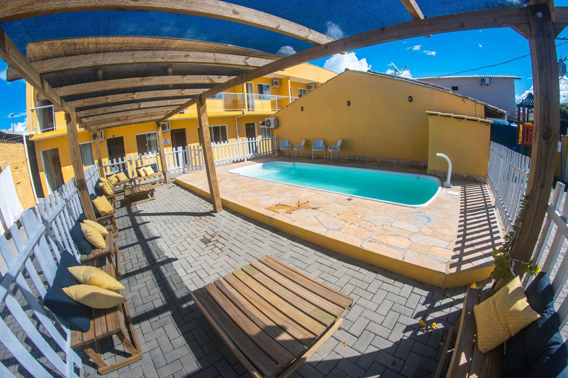 Piscina ensolarada no pátio, com espreguiçadeiras e pérgola de madeira, em um prédio amarelo de um resort.