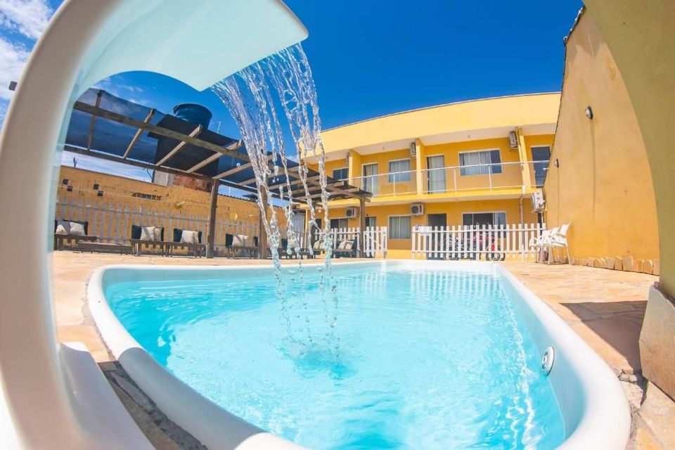 Piscina azul brilhante com cascata em um pátio ensolarado de um resort, com prédios em tons de bege e espreguiçadeiras.