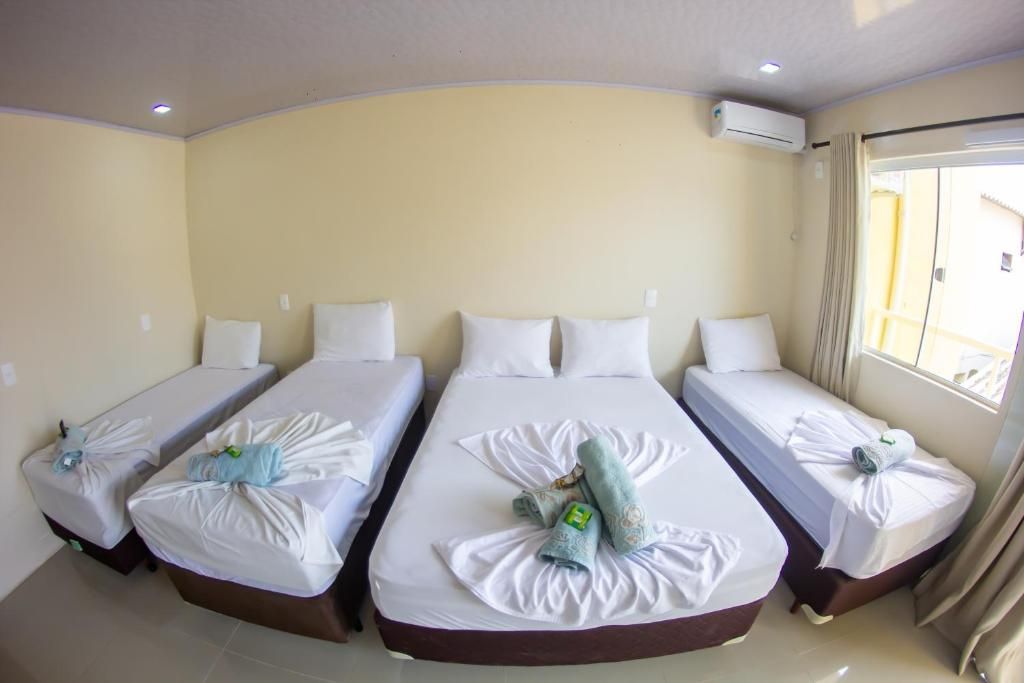 Quarto pequeno com quatro camas de solteiro, lençóis brancos e toalhas enfileiradas perto de uma janela.