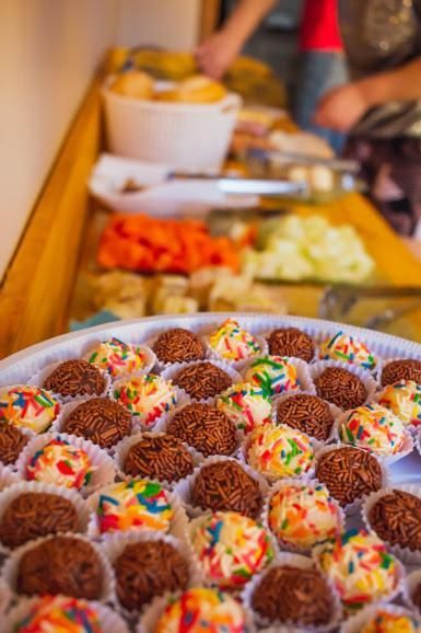 Bandeja de mini donuts com chocolate e granulado em uma barraca de mercado, com frutas desfocadas ao fundo.
