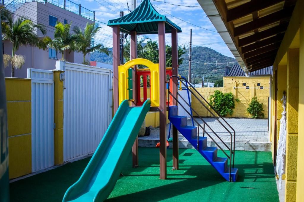 Parque infantil com escorregador verde, escadas azuis e área de recreação sombreada ao lado de uma cerca.