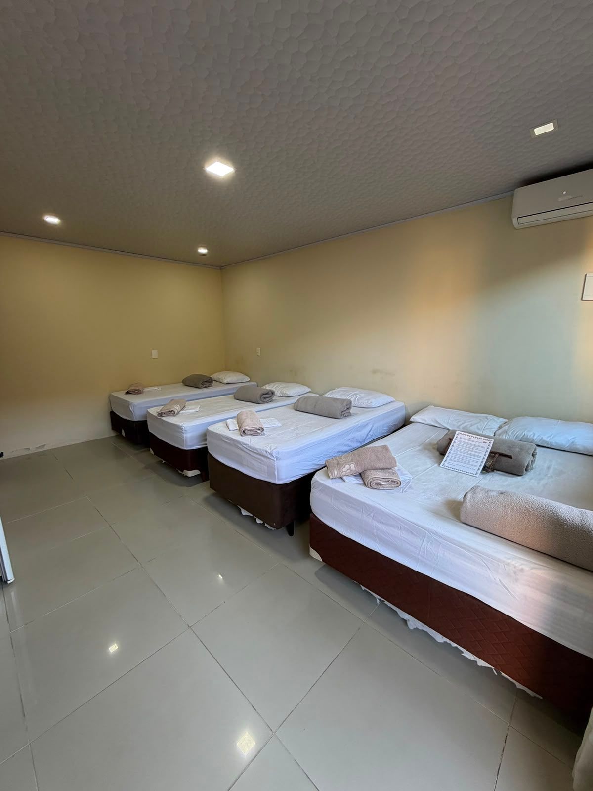Quarto de hotel com quatro camas impecavelmente arrumadas, paredes bege e piso de azulejo brilhante.