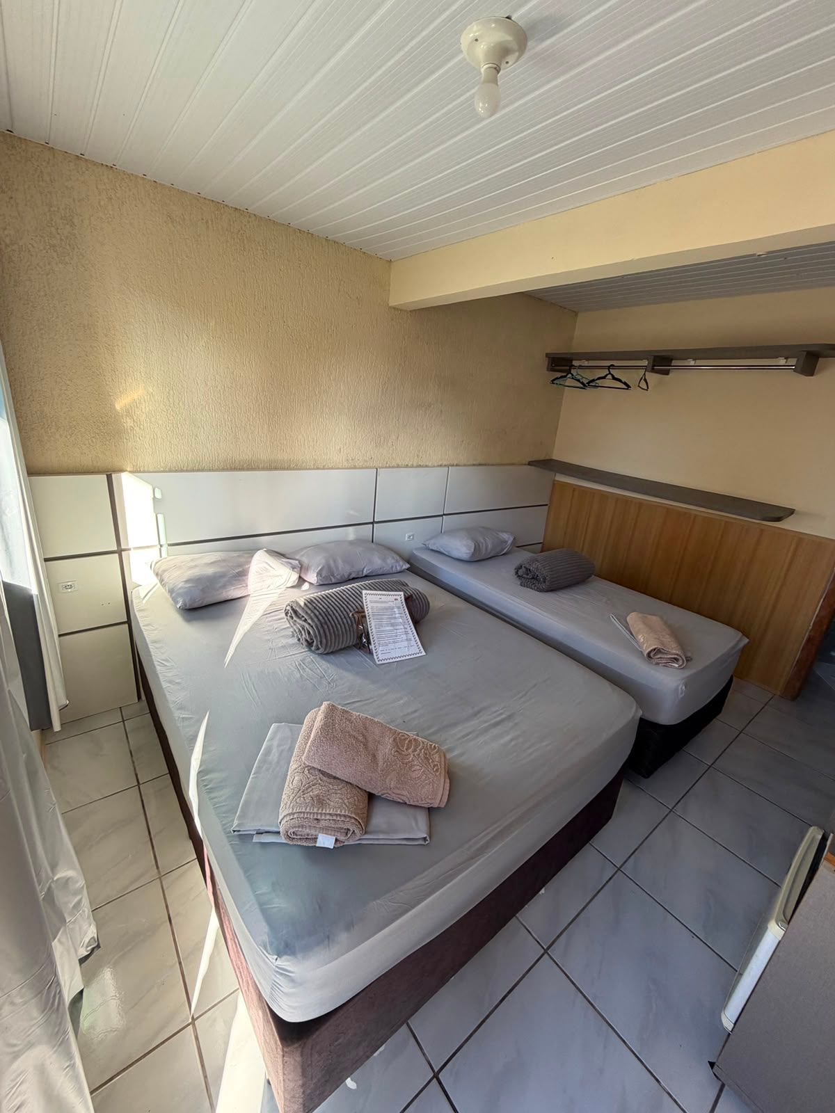 Duas camas de solteiro em um pequeno quarto de hotel bege com piso de azulejos brancos e uma prateleira fixada na parede.