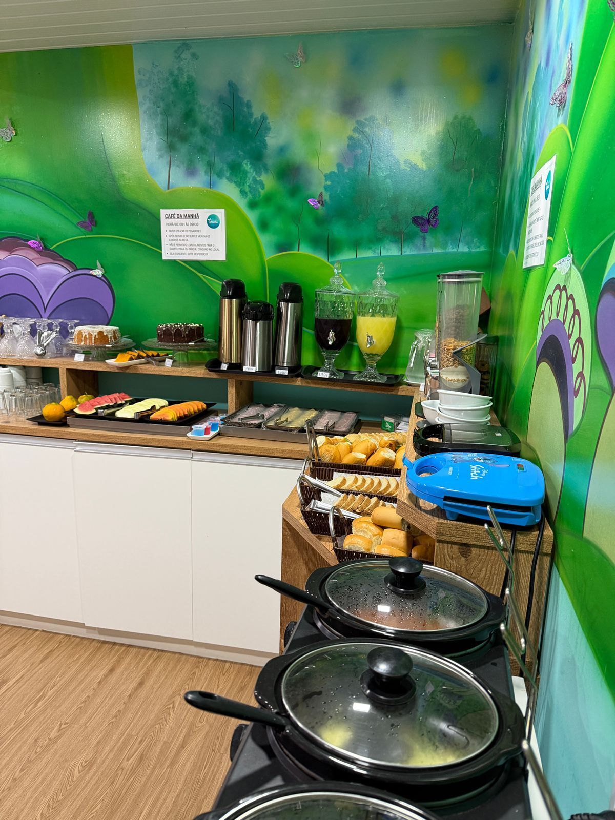 Buffet de café da manhã com cafeteiras, frutas, doces e panelas tampadas ao lado de uma parede com um mural verde.