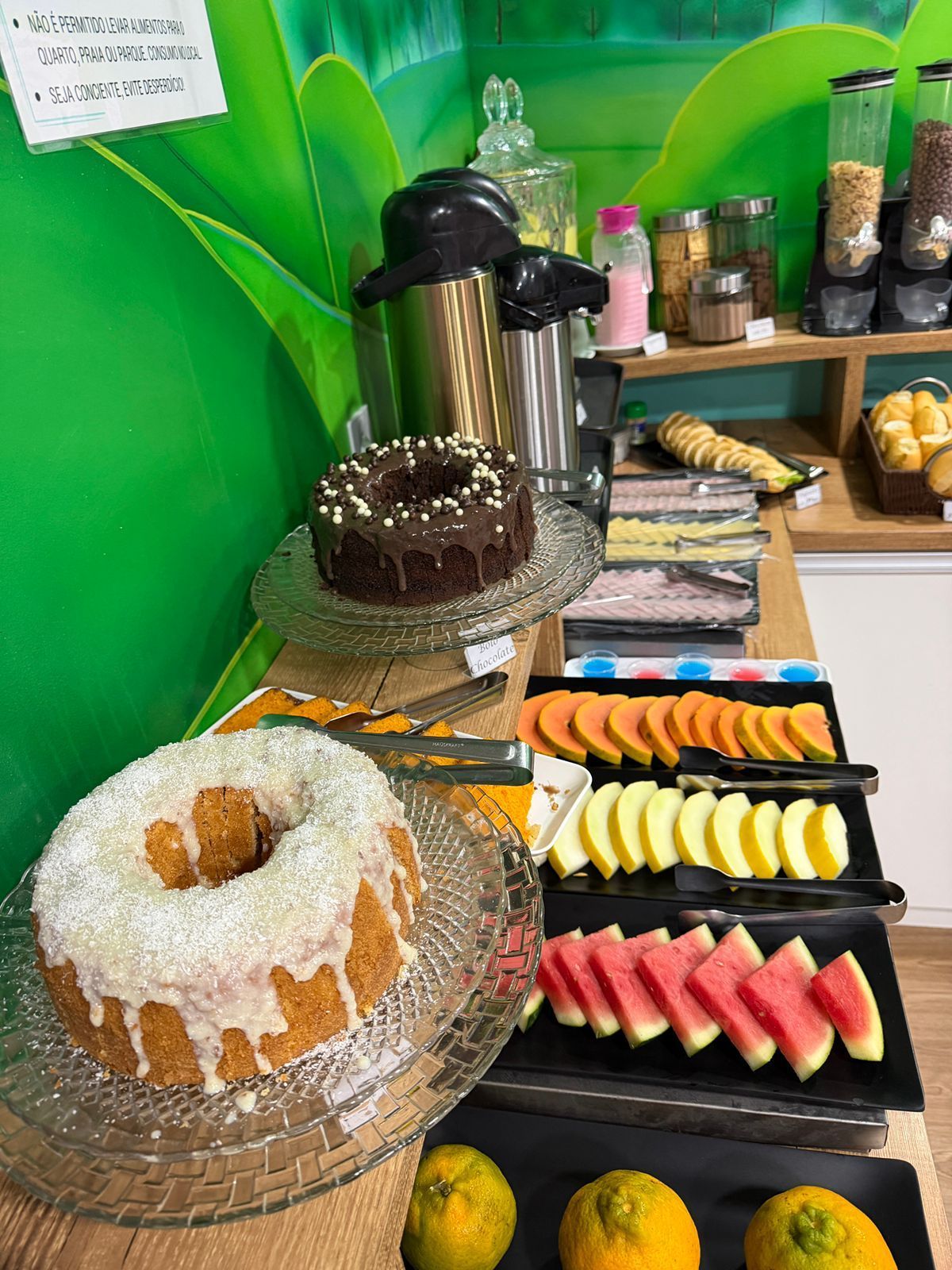 Buffet de sobremesas com bolos, doces, tortas de frutas e máquinas de café em um fundo verde.