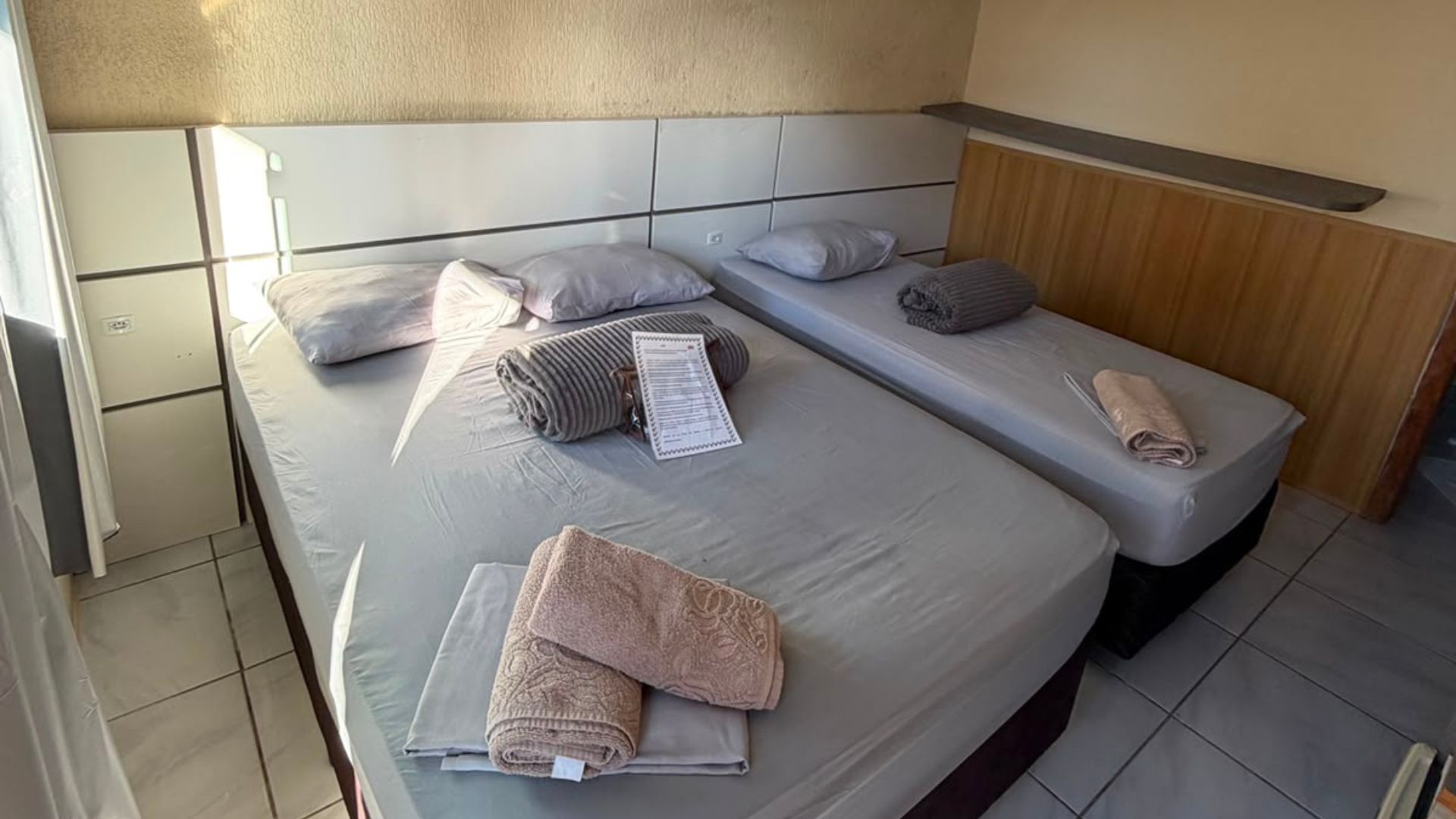Duas camas de solteiro bem arrumadas em um quarto de hotel iluminado, com toalhas dobradas e piso de azulejo.