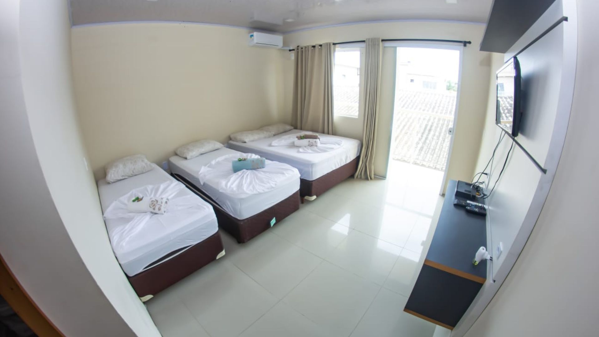 Quarto de hotel pequeno com três camas de solteiro, uma TV de parede e portas de varanda bem iluminadas.