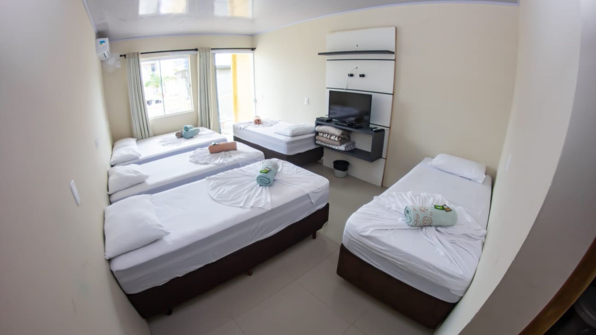 Quarto de hotel pequeno com três camas de solteiro, lençóis brancos, TV, escrivaninha e janela com cortinas.
