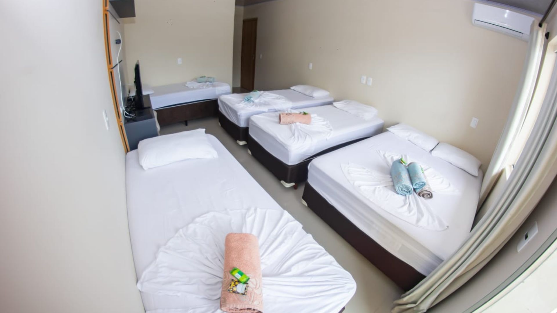 Quarto pequeno com várias camas de solteiro arrumadas e toalhas dobradas, visto da porta.