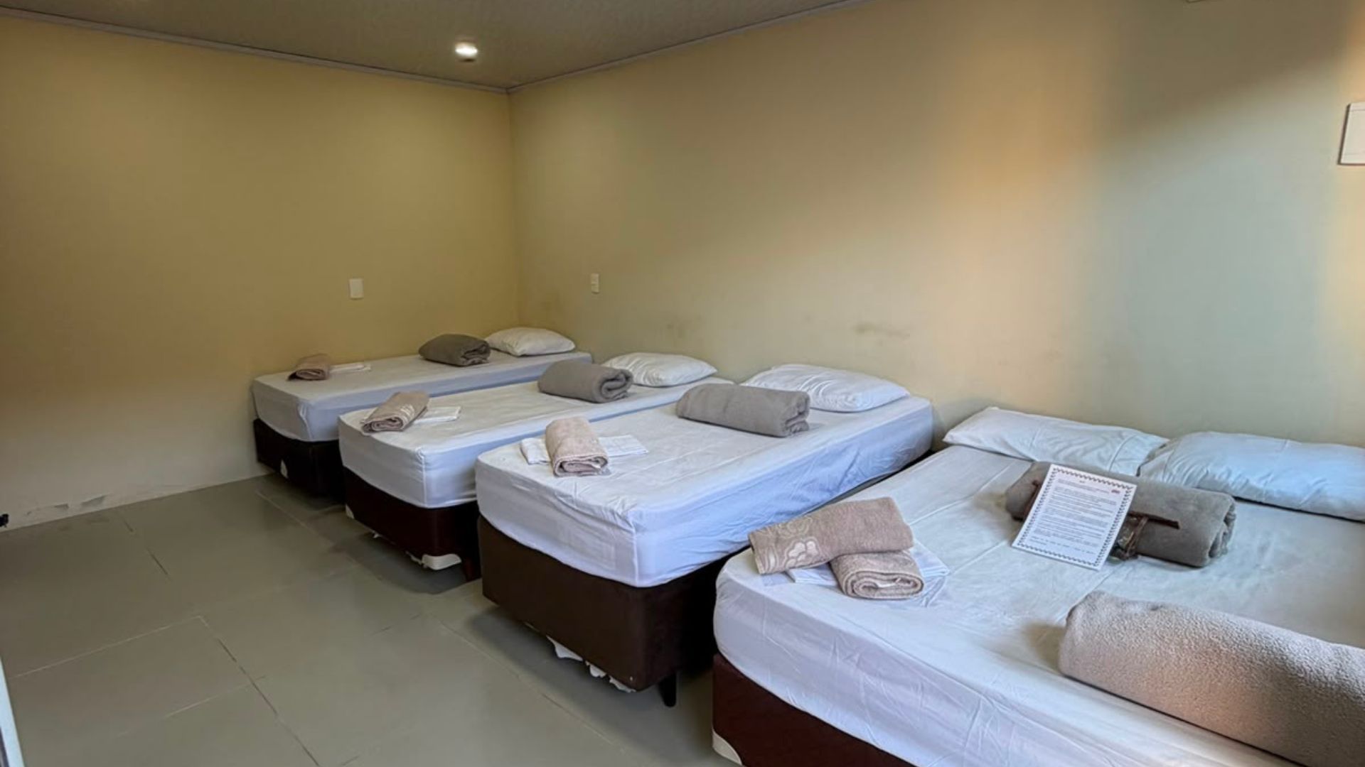 Três camas arrumadas num quarto simples em tons de bege, com toalhas dobradas por cima.