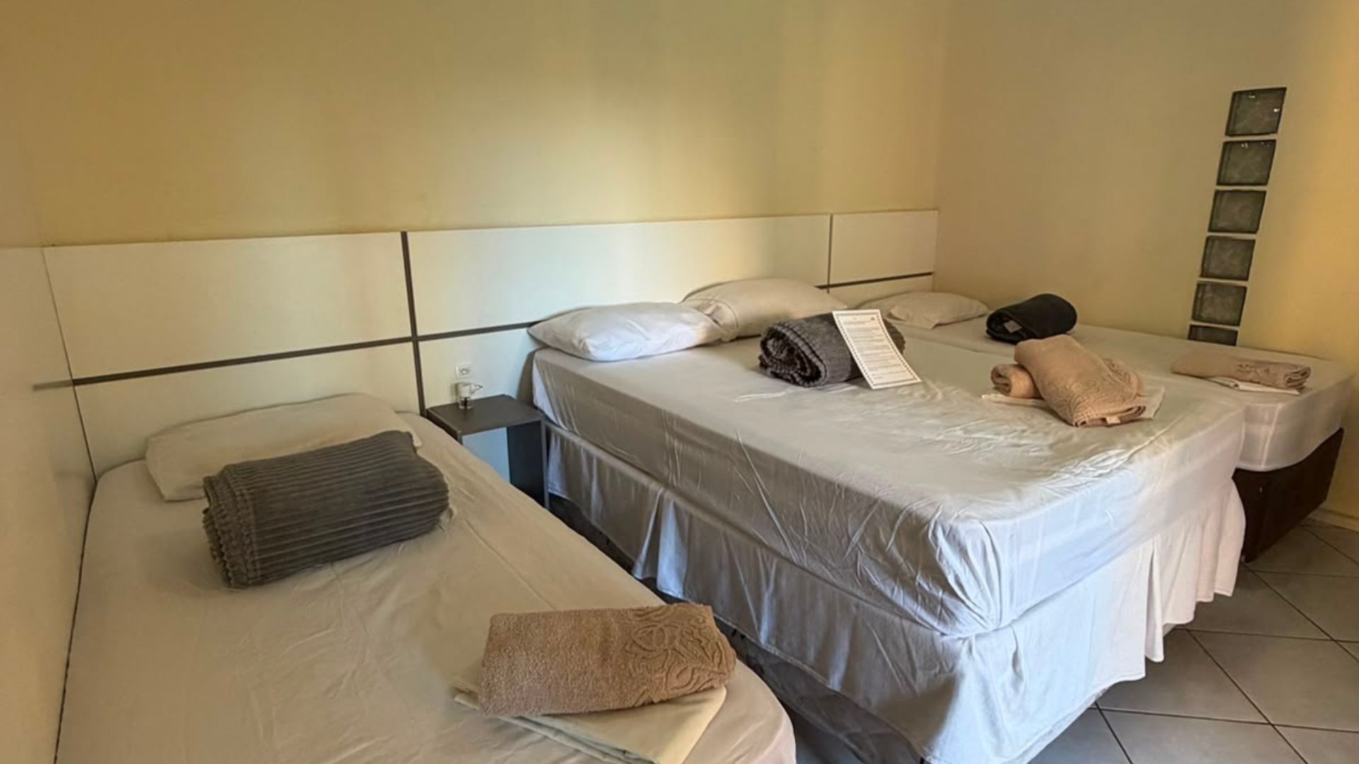 Duas camas arrumadas num quarto de hotel simples, bege, com piso de azulejo e travesseiros.