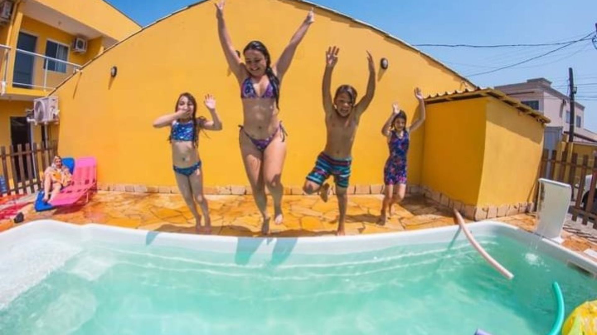 Pessoas pulando em uma piscina em frente a uma casa amarela em um dia ensolarado.