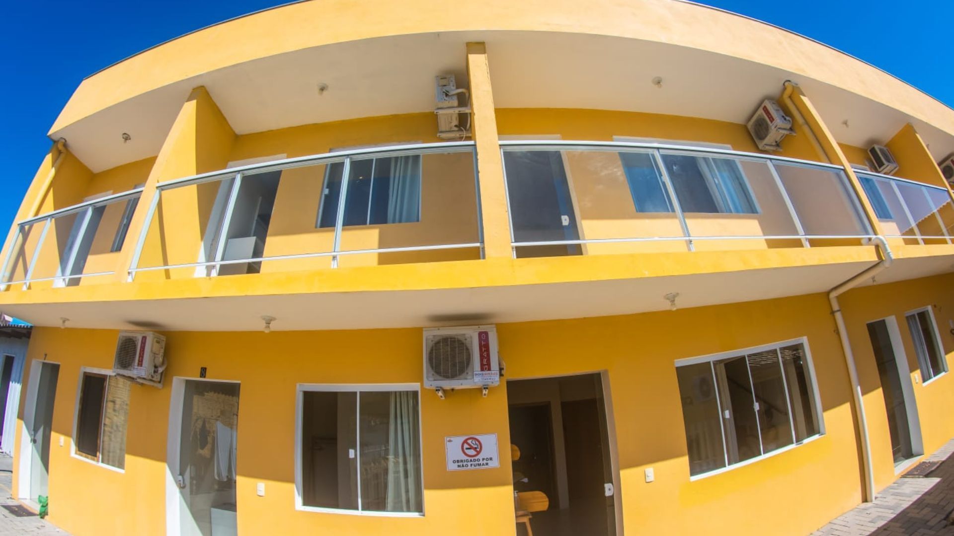 Prédio de apartamentos amarelo de dois andares com varandas, janelas e ar-condicionado, sob forte luz solar.