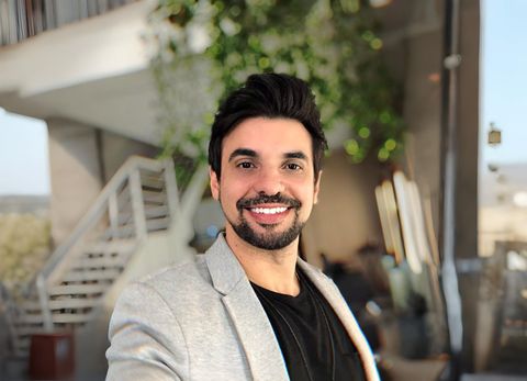 Um homem sorridente de blazer está do lado de fora, perto de escadas e vegetação.
