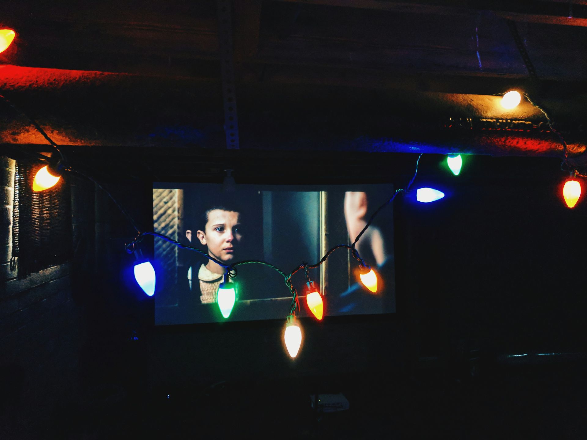 Millie Bobby Brown em uma tela em uma sala escura, com luzes coloridas penduradas na frente.