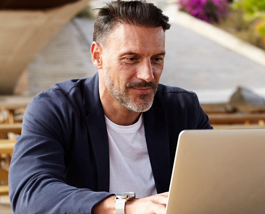 Homem de blazer azul usando um laptop ao ar livre, sorrindo.