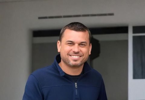 Homem com cabelo castanho curto e barba por fazer sorrindo em frente a um prédio, vestindo um moletom azul.