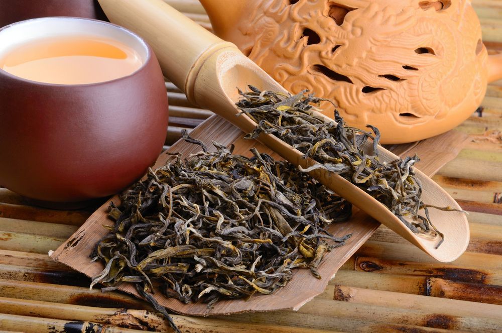 Tea leaves on a bamboo tray beside a clay cup and woven teapot on a rustic table