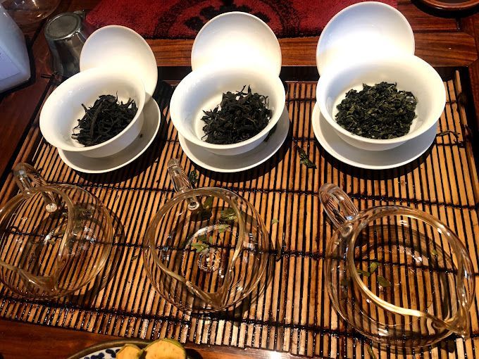Three white bowls of loose tea leaves on a bamboo mat with glass cups and saucers nearby