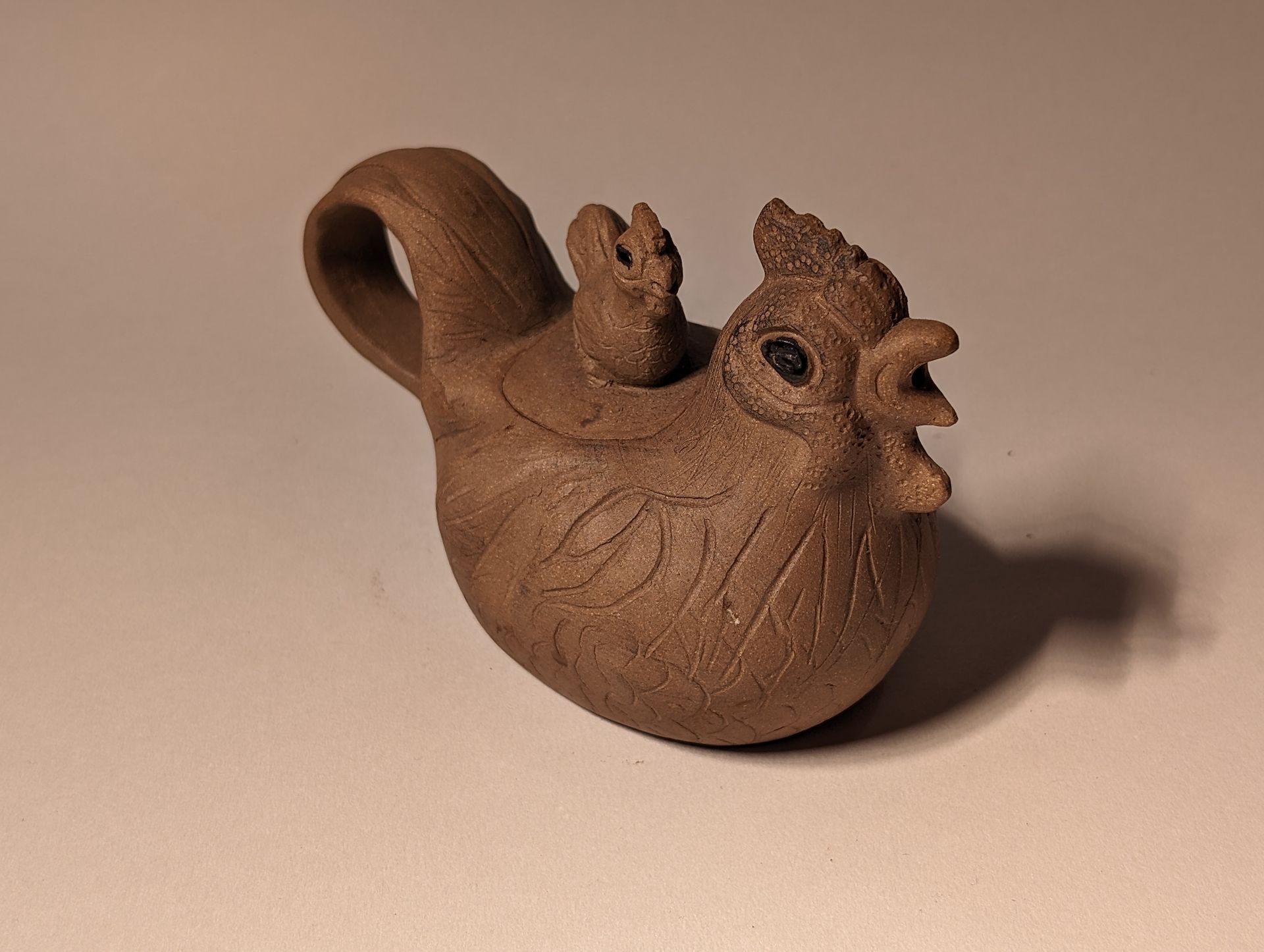 Brown clay figurine of a rooster with a curled tail and open beak on a plain surface