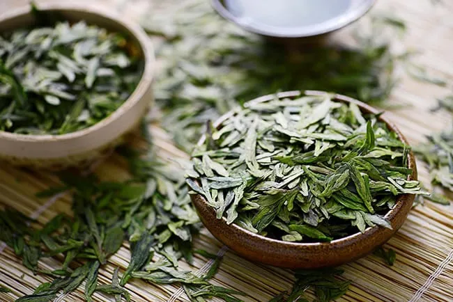 Bowls of loose green tea leaves on a woven mat, with one bowl in focus and others blurred behind.