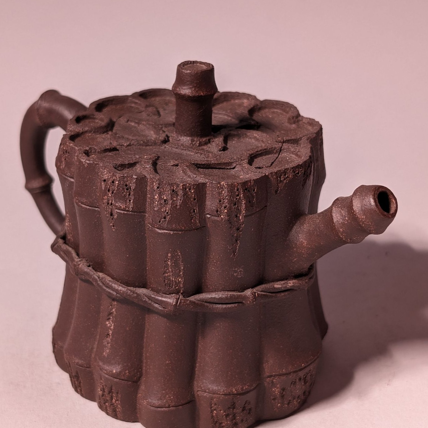 Small brown ceramic teapot with a handle, spout, and decorative top on a plain pink background
