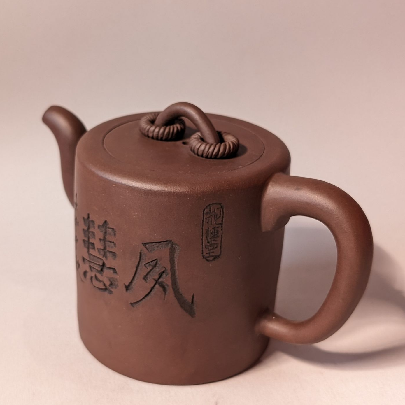 Brown ceramic teapot with Chinese characters and a woven handle on a plain background