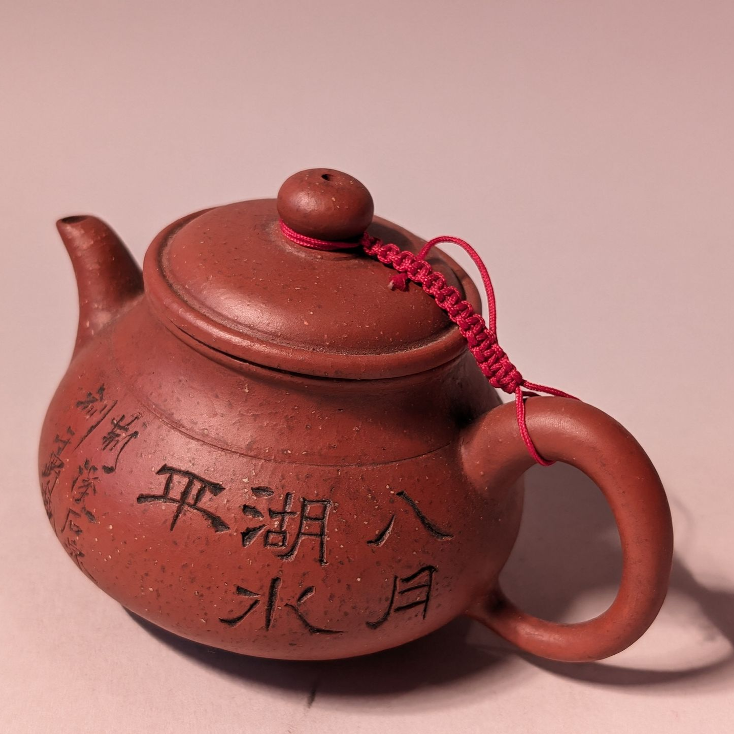 Small reddish-brown teapot with Chinese calligraphy and a red cord on a plain pink background