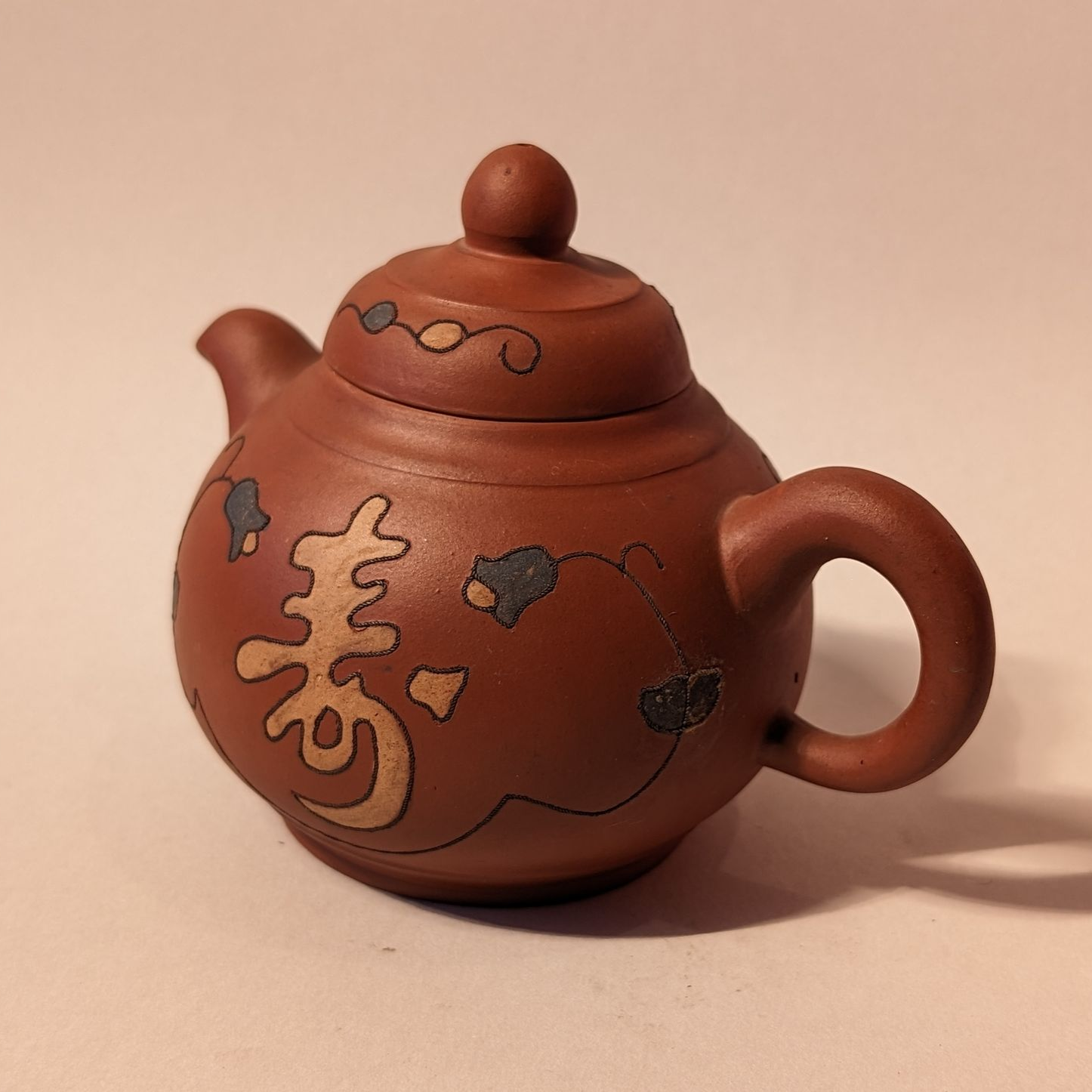 Brown clay teapot with a round lid, decorative patterns, and a curved handle on a beige background