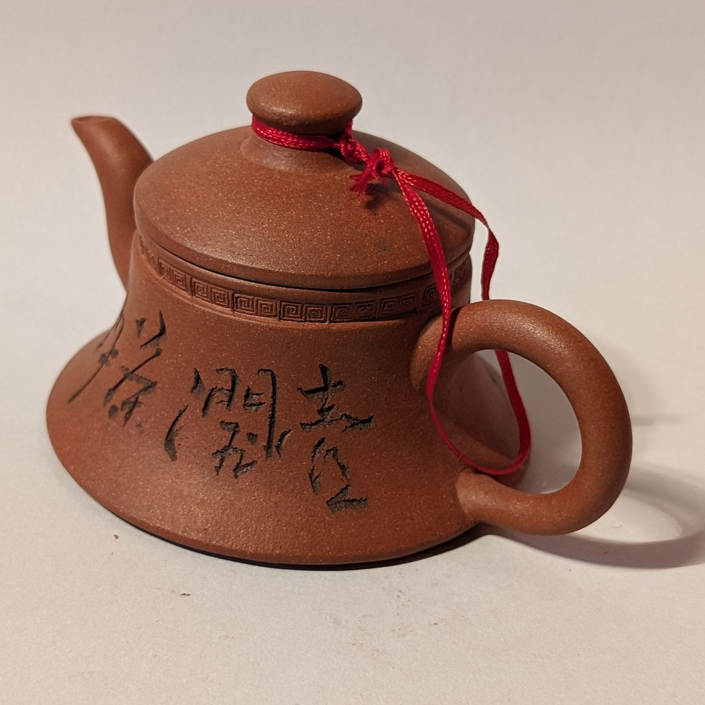 Small brown teapot with a red string and black Chinese characters on a plain background