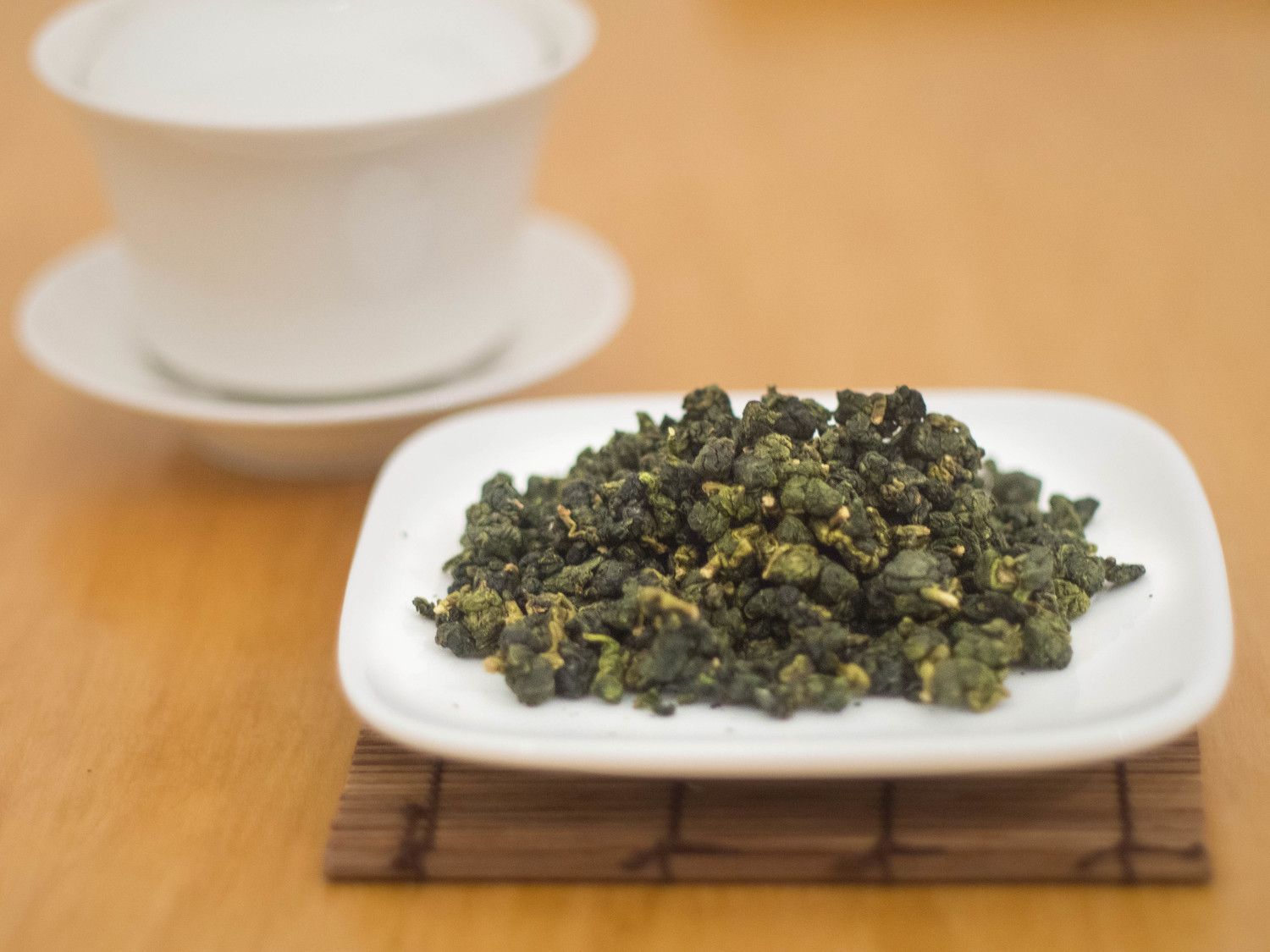 Loose green tea leaves on a white plate beside a white teacup and saucer