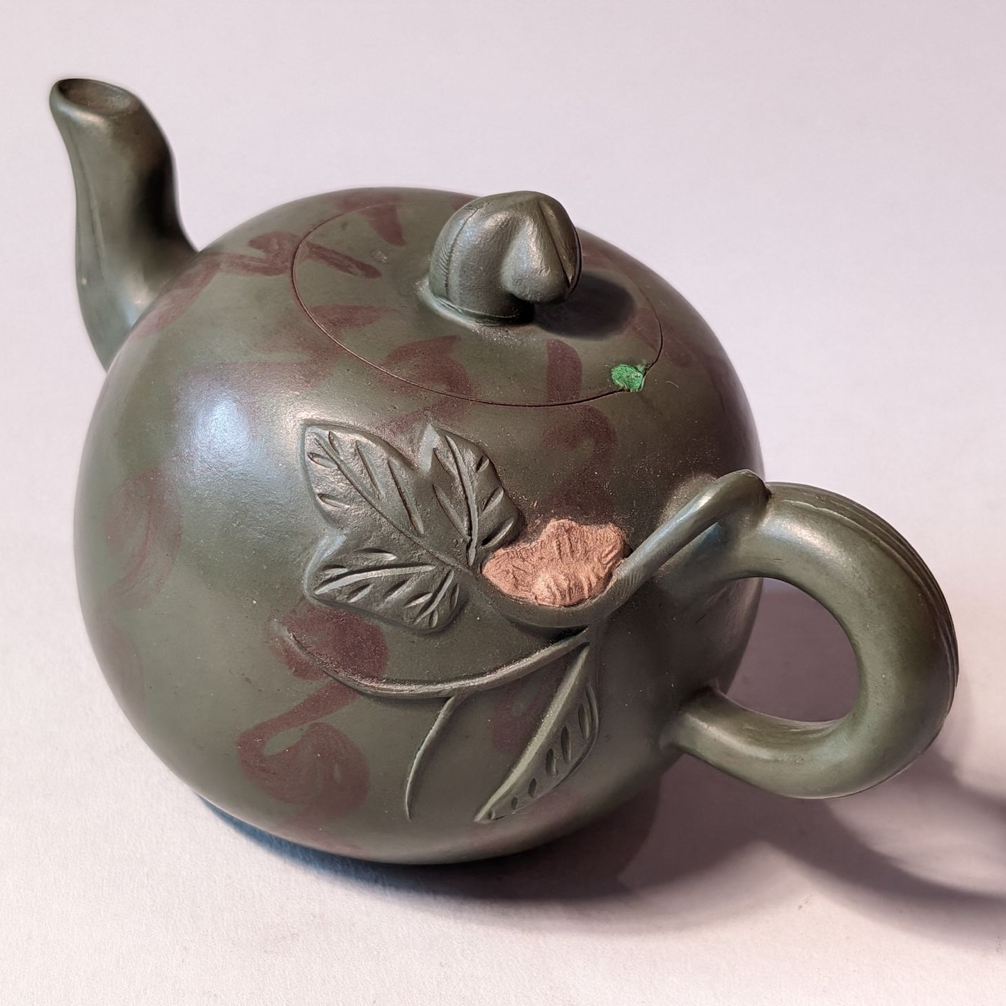 Small green ceramic teapot with leaf decoration and a round body on a light background