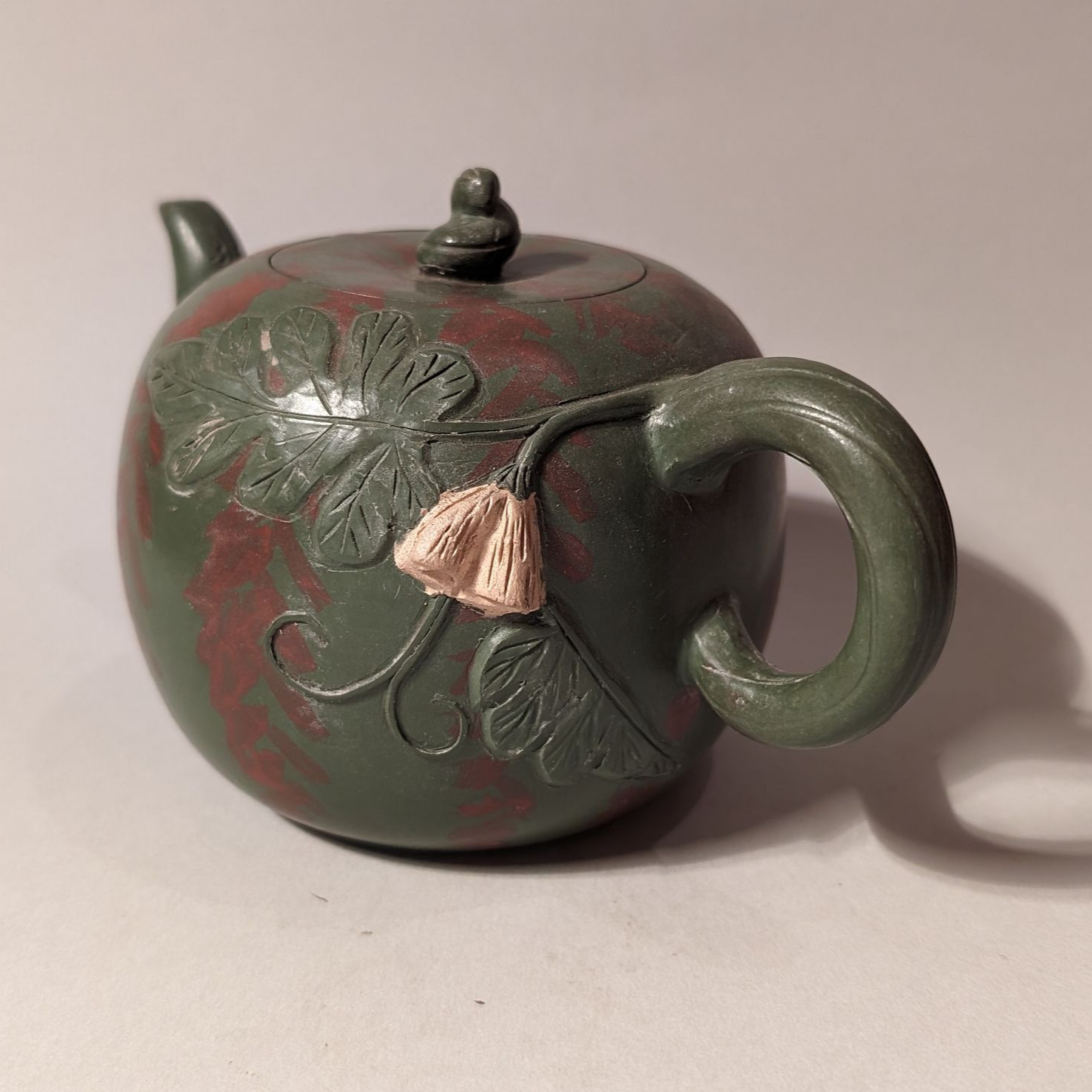 Small green-and-red teapot with a curved handle and floral relief, lit on a plain background