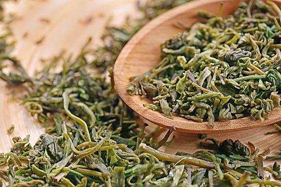 Dried green tea leaves in a wooden bowl with loose leaves scattered around