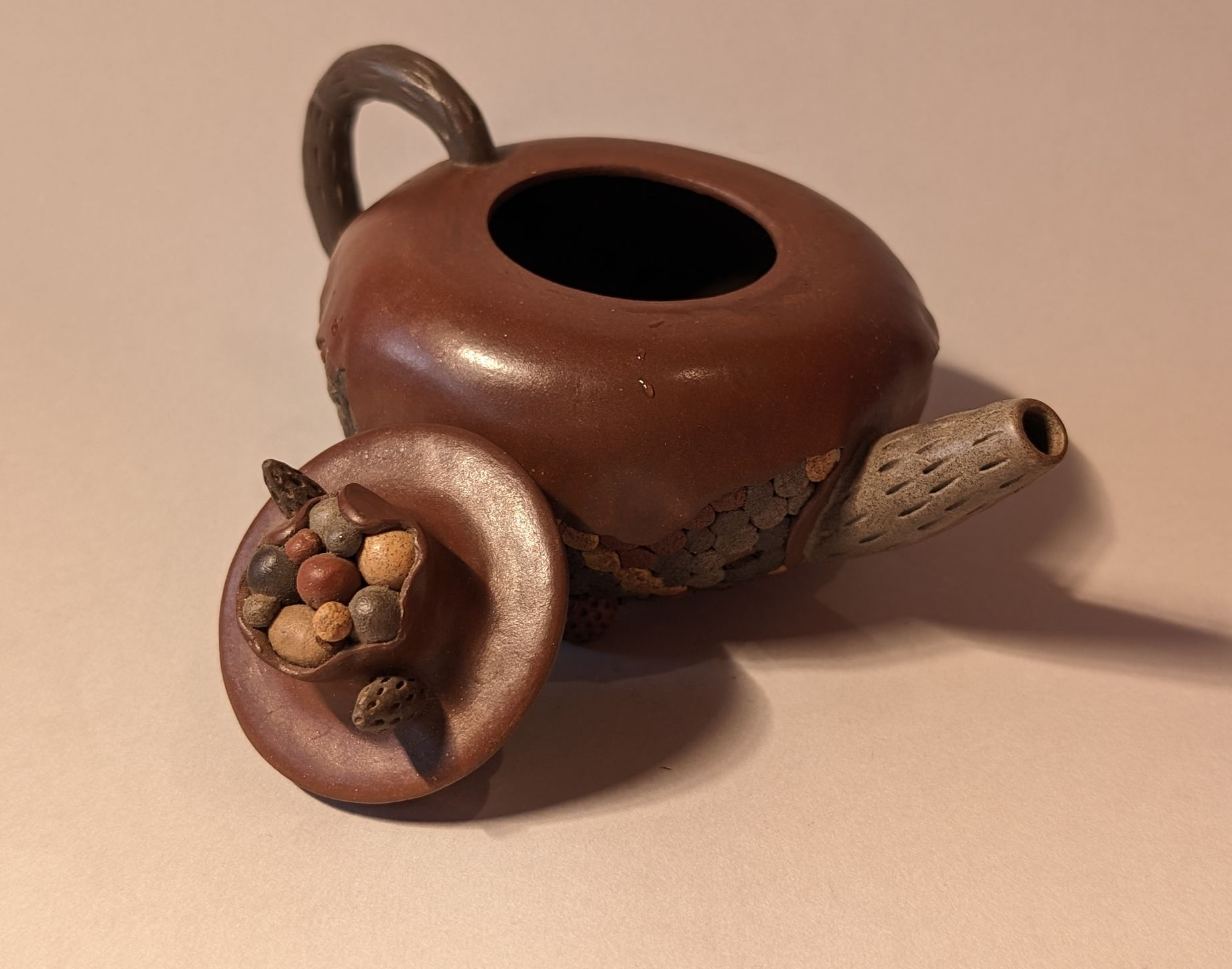 Small brown ceramic teapot with a round opening, lying tipped on its side with a cork stopper and tea leaves nearby