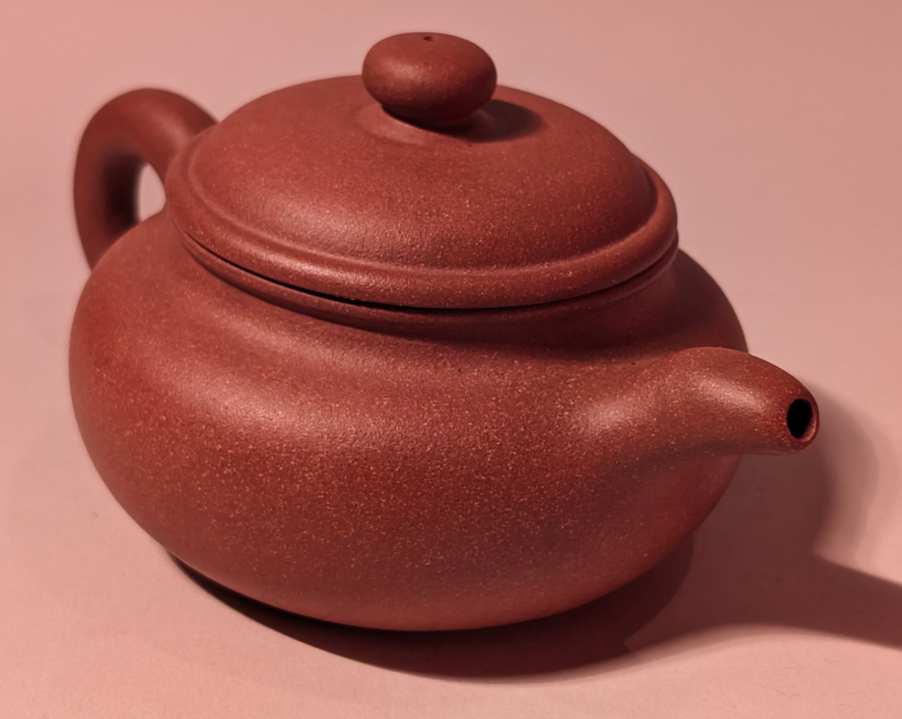 Red ceramic teapot with lid and side handle on a pink surface